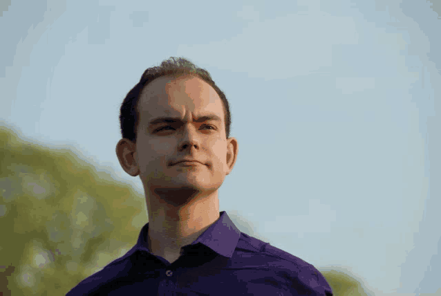 a man in a purple shirt looks at the camera with trees in the background