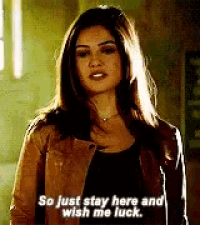 a woman in a brown jacket is standing in a dark room and saying `` so just stay here and wish me luck '' .