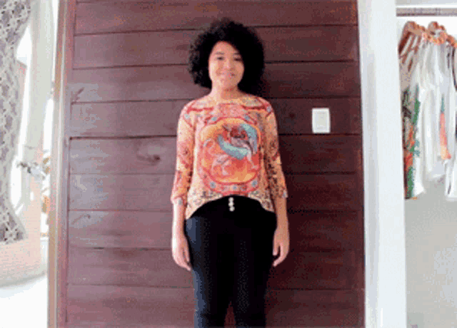 a woman standing in front of a wooden wall wearing a colorful shirt