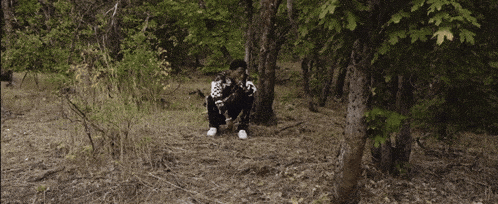 a man in a black and white jacket sits on the ground in the woods
