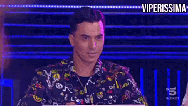 a man in a colorful shirt is sitting in front of a microphone and a sign that says viperissima .