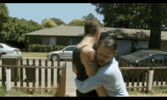 two men hugging in front of a house with cars parked in the driveway