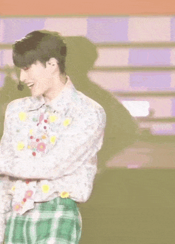 a young man in a floral shirt and green plaid skirt is standing on a stage with his arms crossed .