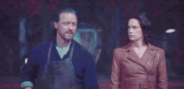 a man and a woman are standing next to each other in a dark room . the woman is wearing a brown leather jacket .