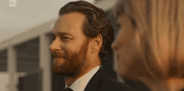 a man with a beard is smiling in front of a woman with rai written on the bottom of the screen