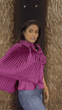 a woman wearing a purple shirt and blue jeans stands in front of a wall