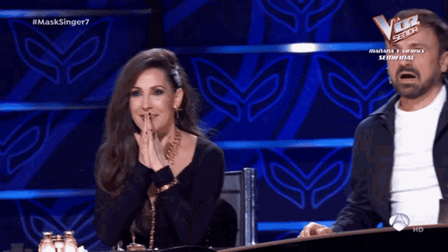 a man and a woman are sitting at a table in front of a screen that says # masksinger7