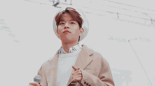 a young man in a tan coat and white hat is holding a microphone and giving a thumbs up