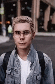a young man wearing glasses and a denim jacket is walking down a street .