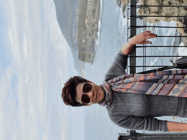 a woman wearing sunglasses and a scarf leans against a fence overlooking the ocean