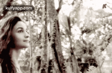 a black and white photo of a woman standing in the woods looking up at the sky .