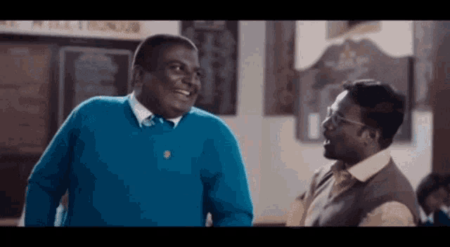 two men are laughing in a room and one of them is wearing a blue sweater .