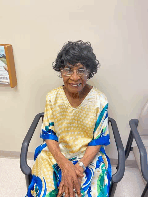 an elderly woman wearing glasses sits in a chair with her hands folded