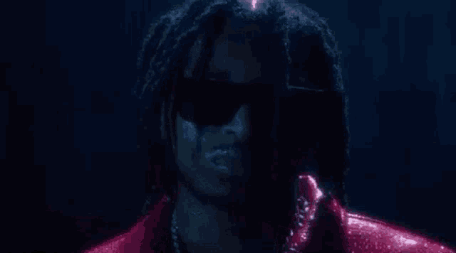 a man with dreadlocks wearing sunglasses and a red jacket is standing in the rain .