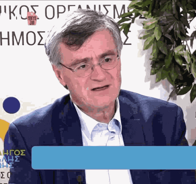 a man wearing glasses and a suit is sitting in front of a sign that says ' kos organism '