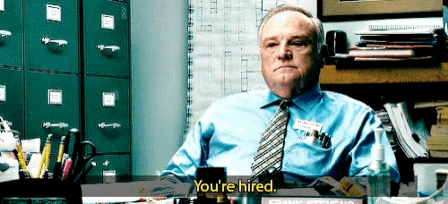 a man in a blue shirt and tie is sitting at a desk and says " you 're hired "