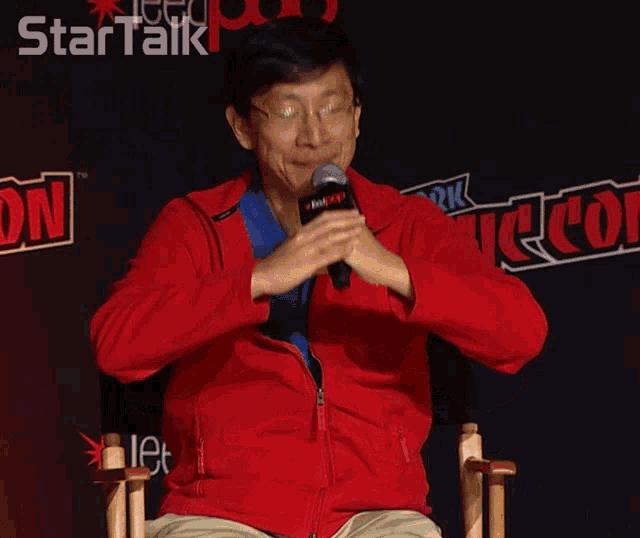 a man in a red jacket holds a microphone in front of a sign that says comic con