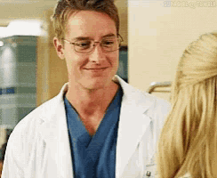 a man in a lab coat and scrubs is talking to a woman in a hospital room .