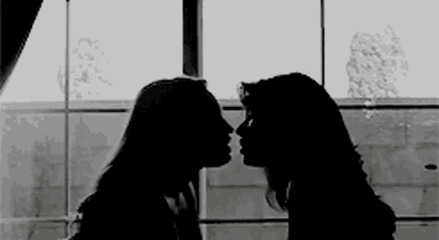 two women are kissing in front of a window .