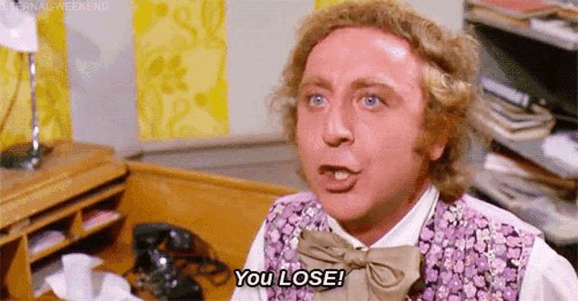 a man in a purple vest and bow tie is saying `` you lose '' while sitting in front of a desk .