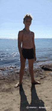a shirtless young man stands on a sandy beach in front of the ocean ..
