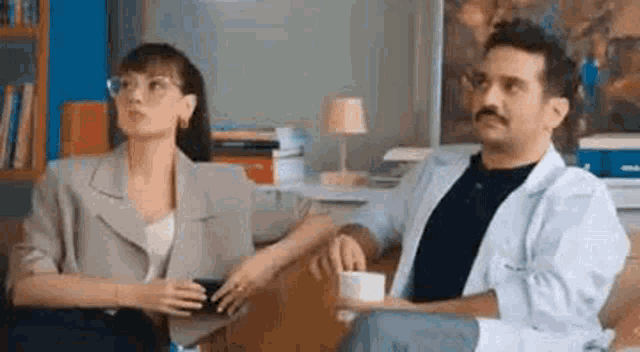 a man and a woman are sitting next to each other on a couch . the woman is holding a cup of coffee .