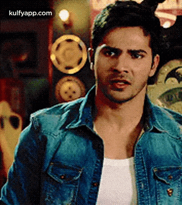 a man wearing a denim jacket and a white tank top is standing in front of a wall with plates on it .