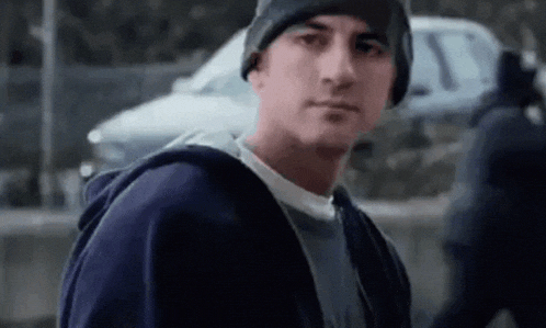 a man wearing a beanie and a hoodie is standing in front of a car and looking at the camera .