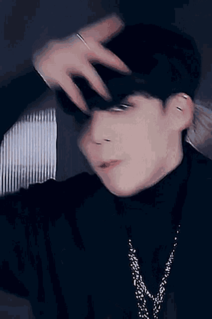 a young man wearing a black turtleneck and a necklace holds his hand to his face