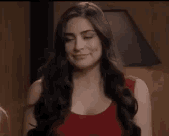 a woman with long dark hair is wearing a red tank top and smiling with her eyes closed .