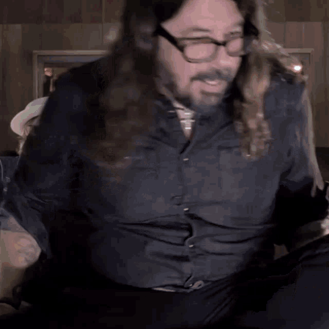 a man with long hair and glasses sits on a couch with his legs crossed