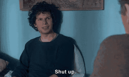 a man with curly hair is sitting in a chair talking to another man and saying `` shut up '' .