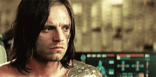 a man with long hair and a bandage on his arm is standing in front of a computer screen .