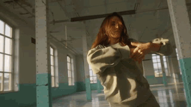 a woman in a grey sweatshirt is dancing in a room with many windows