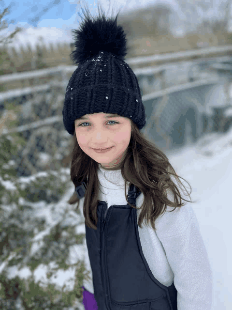 a little girl wearing a black beanie and overalls stands in the snow