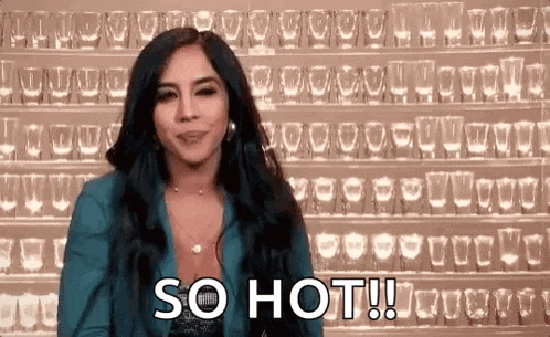 a woman is standing in front of a wall of shot glasses and saying `` so hot ! ''