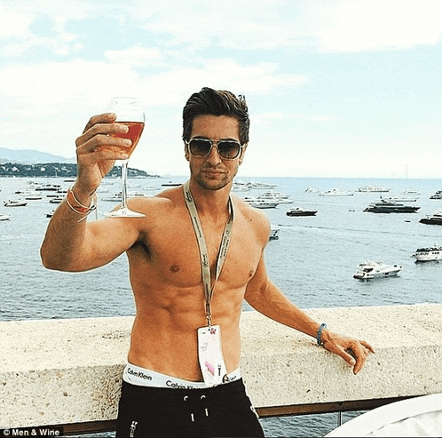a shirtless man holding a glass of wine in front of the ocean