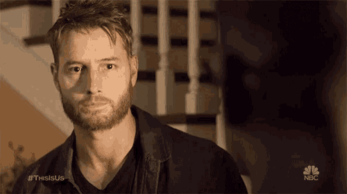 a man with a beard is standing in front of a staircase and looking at another man .