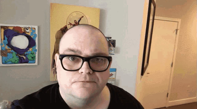 a bald man wearing glasses stands in front of a painting on the wall