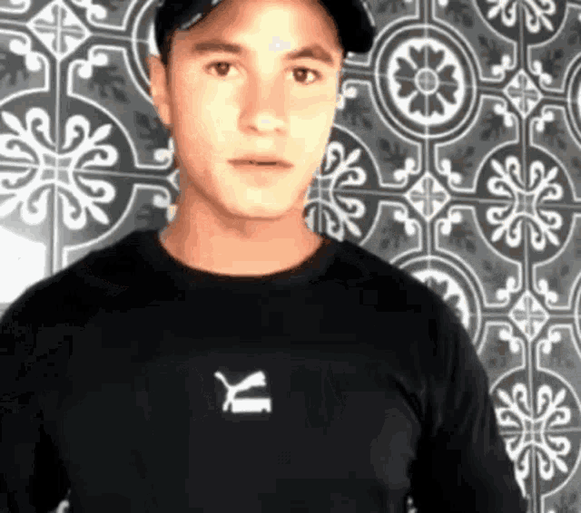 a man wearing a black puma shirt stands in front of a tile wall