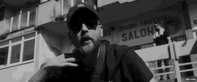 a man with a beard wearing sunglasses and a hat is standing in front of a building that says salon .