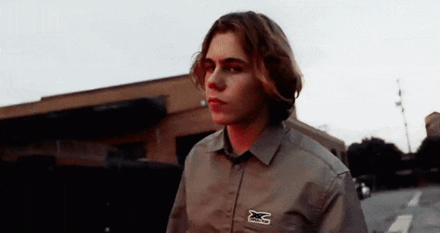 a young man with long hair is standing in front of a building wearing a khaki shirt .