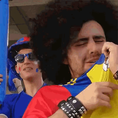 a man wearing a wig and sunglasses talks on a cell phone next to another man