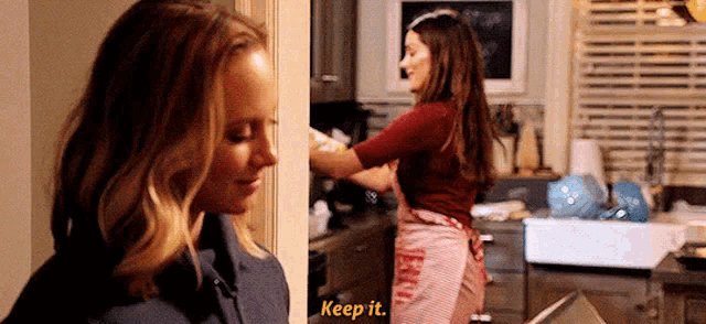 a woman in an apron is standing next to another woman in a kitchen and says `` keep it '' .