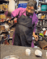 a woman in a pink shirt and black apron is standing in a messy kitchen