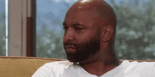 a bald man with a beard is sitting on a couch and making a face .