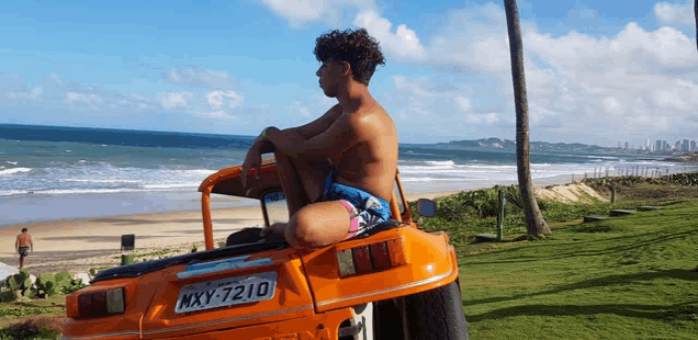 a shirtless man sits in an orange vehicle with a license plate that says mxy 7210