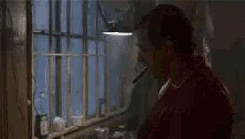 a man in a red shirt is smoking a cigarette in a kitchen .