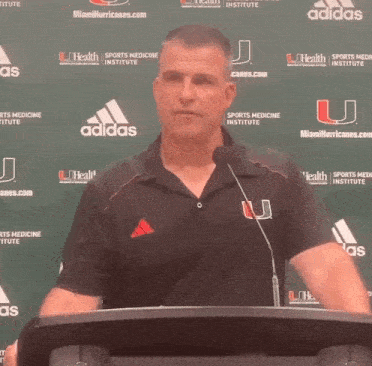 a man stands at a podium in front of a miami hurricanes.com banner