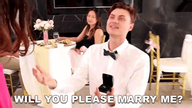 a man in a bow tie is holding a ring in a box and asking a woman to marry him